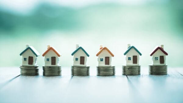 Tiny figurine houses sitting in a row on top of piles of coins.